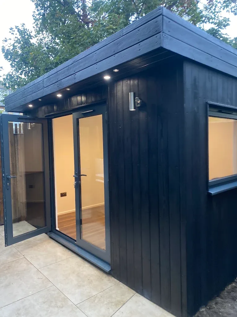 A black-clad garden office at dusk with soffit downlights and glazed doors open to a lit interior.