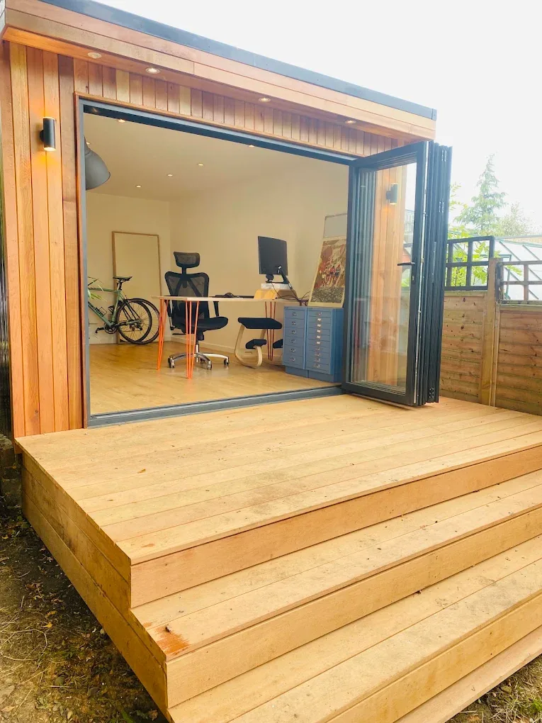 A cedar garden office with bi-fold doors open onto timber deck steps.