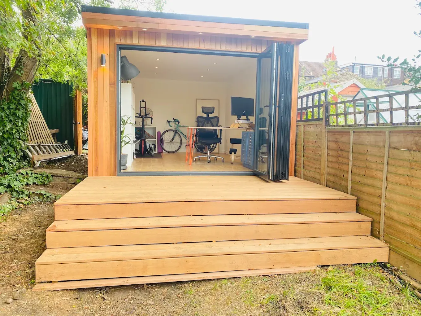 A cedar garden office with bi-fold doors open onto a timber deck, desk and chair inside.